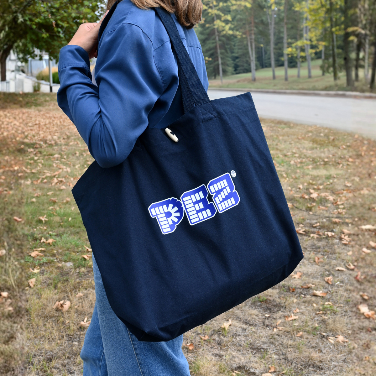 Eine Person, von hinten gesehen, trägt die dunkelblaue PEZ-Baumwolltragetasche über der Schulter auf einem herbstlichen Weg.